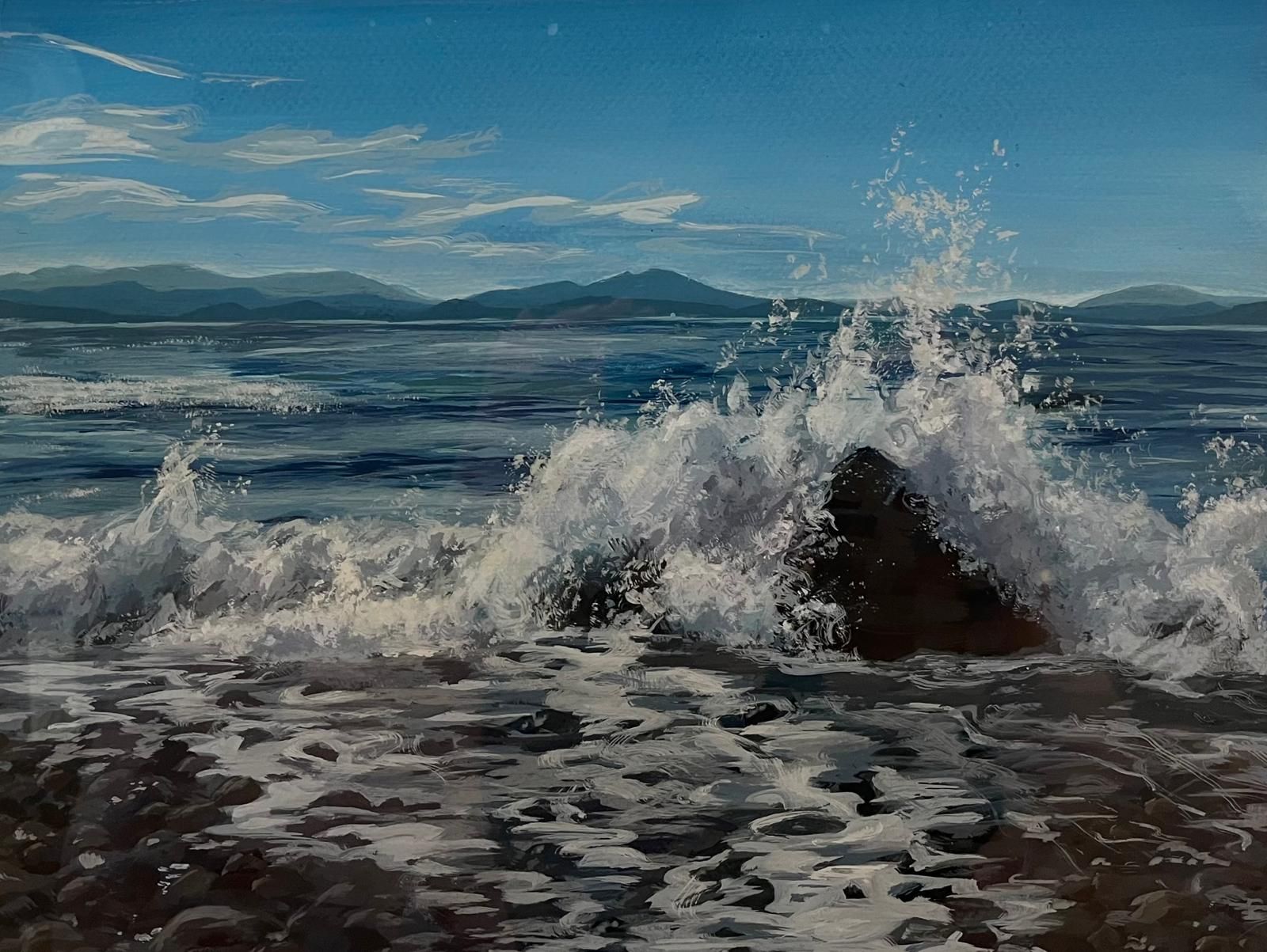 A wave breaking white over dark stones in the foreground, with flat blue water beyond and low blue-green mountains along the far shore.